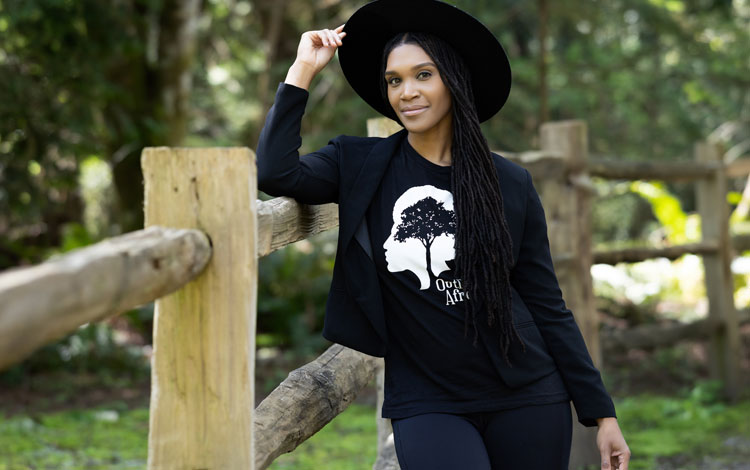Woman wearing a black shirt and a black hat leaning against a fence with her hand on her hat, posing.